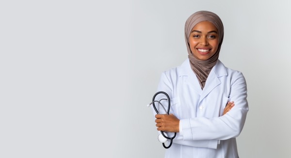A woman standing in front of a grey background with a lab coat and head scarf, arms crossed, holding a stethoscope.